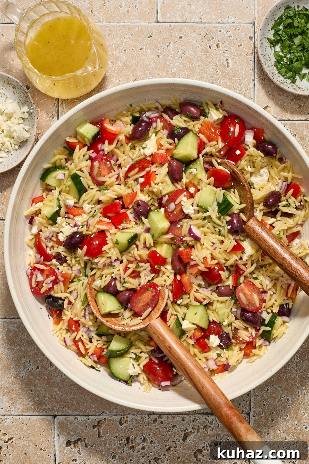 Salad in a large white bowl with wooden servers and chopped parsley, feta and lemon dressing surrounding it.