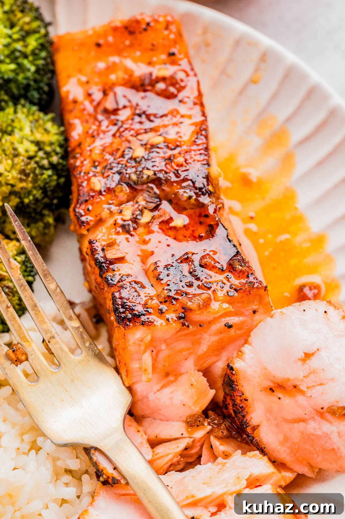 Close-up of a cooked salmon fillet with seasoning and sauce, served with white rice and broccoli on a plate, alongside a fork.