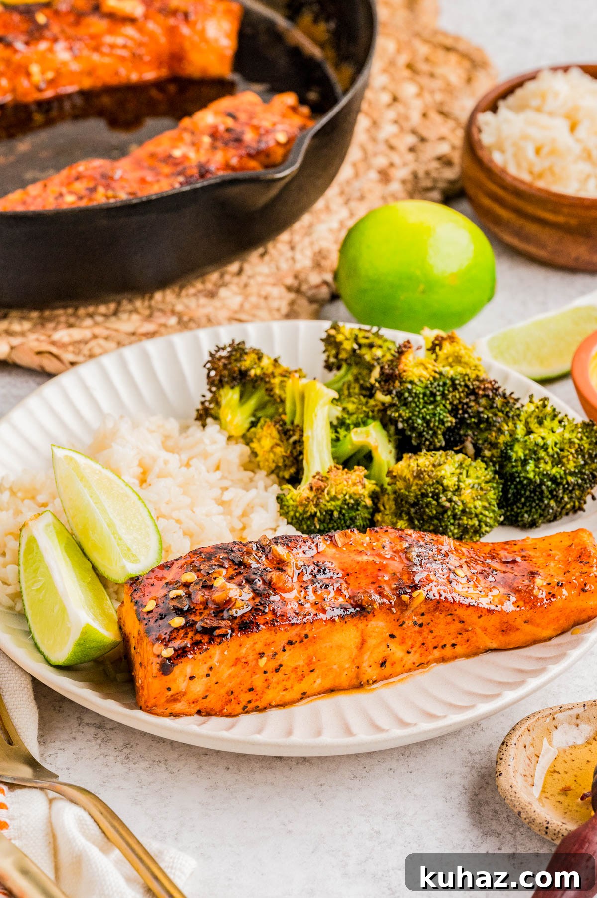 A plate with a cooked salmon fillet, white rice, roasted broccoli, and lime wedges, with a skillet, bowl of rice, and lime in the background.