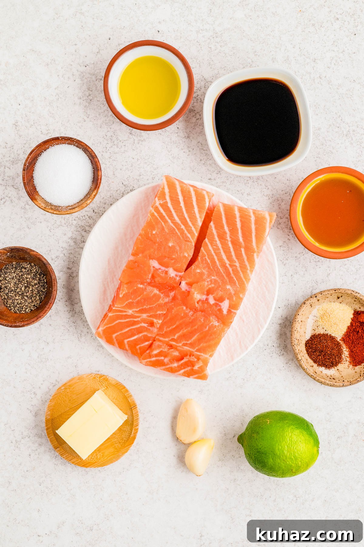 Two raw salmon fillets on a plate, surrounded by small bowls of olive oil, soy sauce, honey, salt, pepper, spices, butter, garlic cloves, and a whole lime on a white surface.