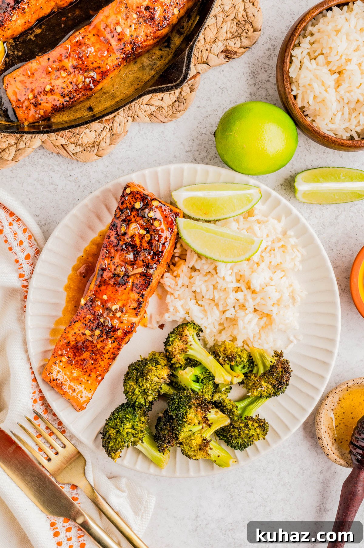 A plate with grilled salmon topped with spices, steamed white rice, roasted broccoli, and lime wedges, with extra rice and a cast iron pan of salmon nearby.