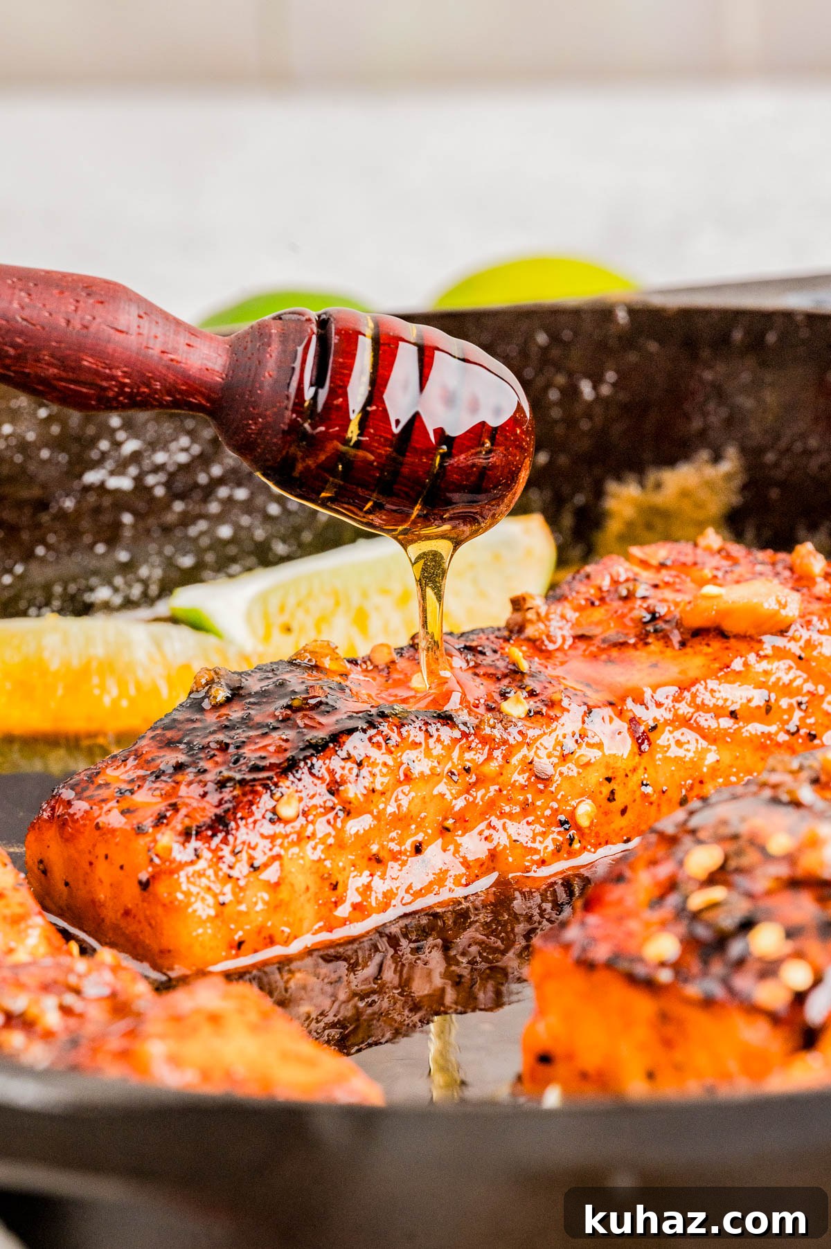 A hand drizzles honey over a seared, seasoned salmon fillet in a skillet, with lime wedges visible in the background.