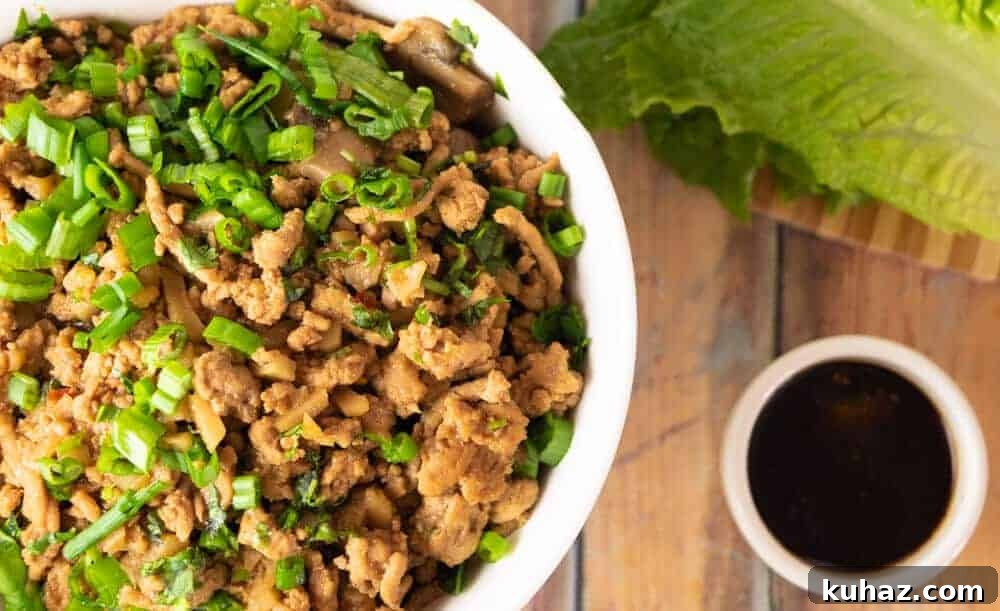 Instant Pot Chicken Lettuce Wraps: Copycat Takeout Recipe 2 Close-up of Instant Pot chicken lettuce wraps filling in a white bowl, topped with fresh chopped scallions, with lettuce leaves and dipping sauce in the background.