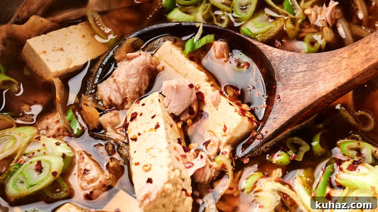 19 Asian Recipes That Fed My Family for $20 15 Close-up of a wooden spoon in a vibrant kimchi stew with tofu and pork, garnished with sliced green onions and red pepper flakes.