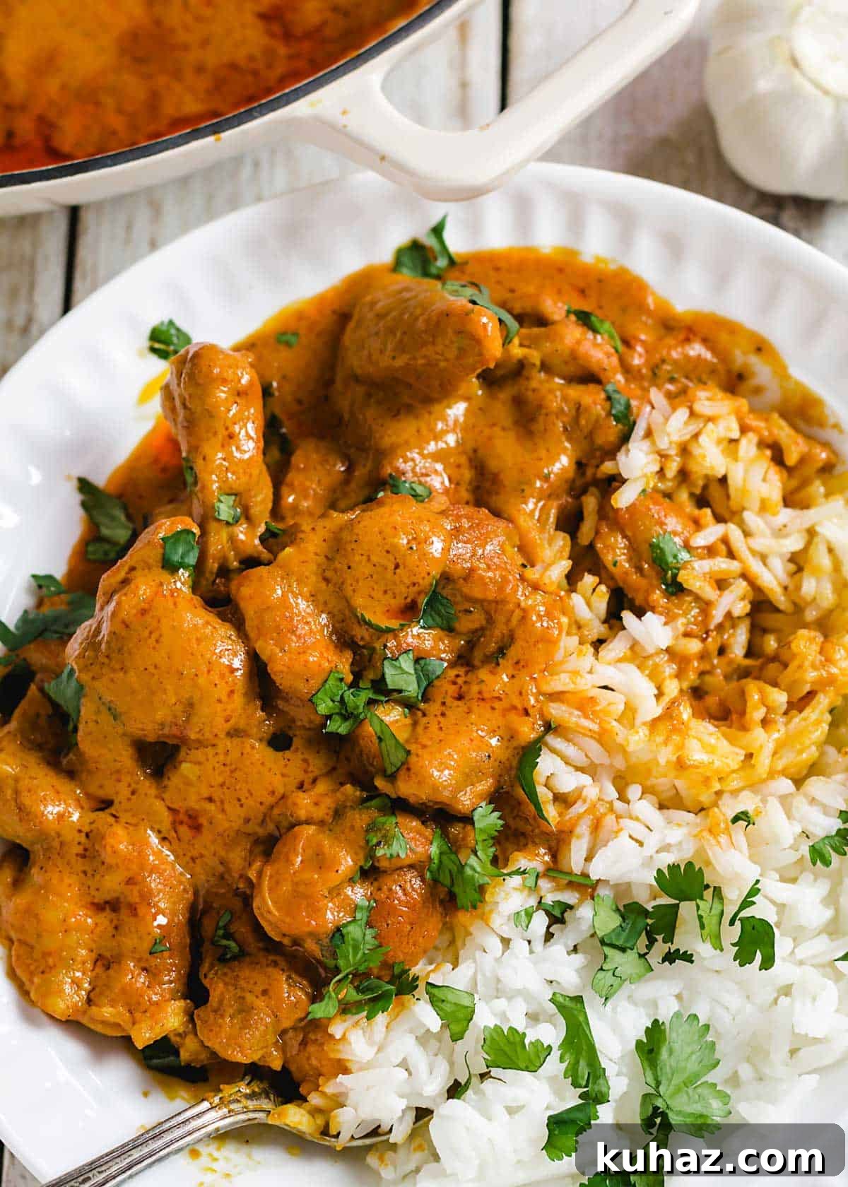 A close up of chicken tikka masala in a bowl next to white rice and garnished with cilantro leaves.
