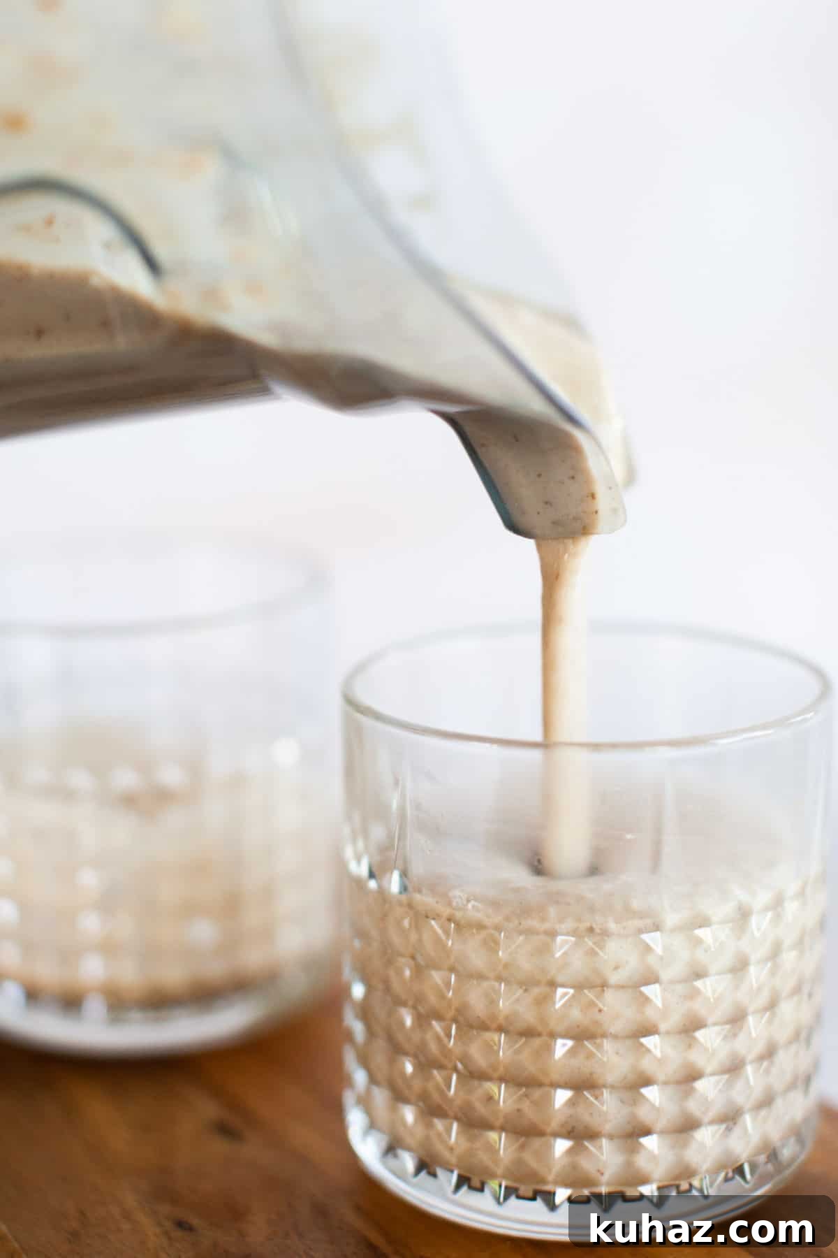 A thick, creamy coffee smoothie is being poured from a blender into a textured glass, with another glass partially visible in the background.