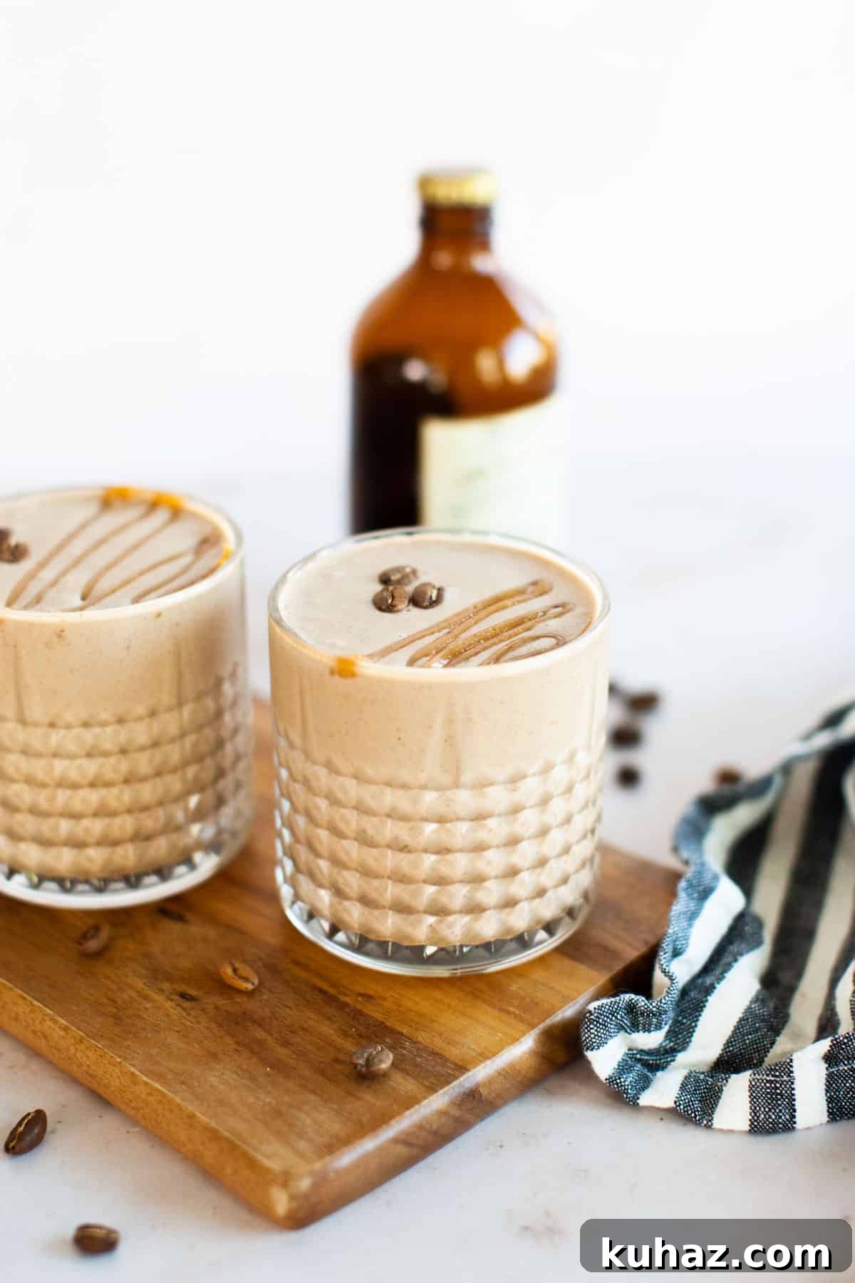 Two glasses of creamy coffee smoothie with a drizzle on top sit beside coffee beans on a wooden board, accompanied by a striped cloth and a brown bottle in the background.
