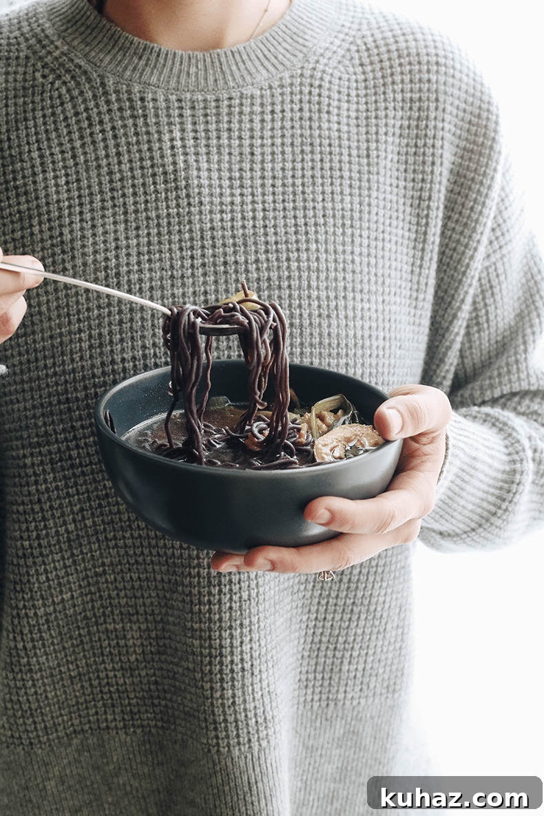 Person in a grey waffle sweater holding a bowl of healing soup. Silver spoon taking a scoop of noodles and vegetables out of the soup.