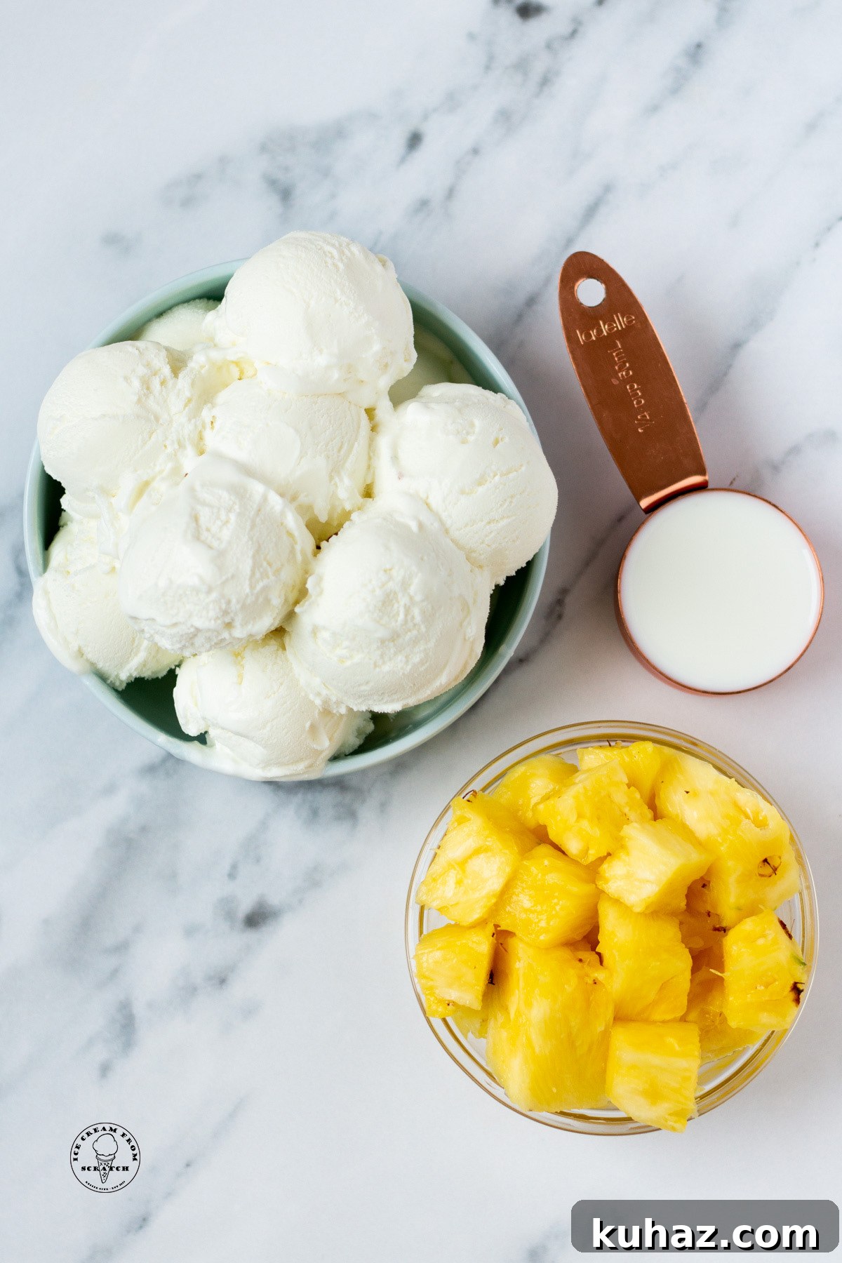 a large bowl of scooped vanilla ice cream, a smaller bowl of diced pineapple, and a measuring cup holding milk, on a marble counter, viewed from above.