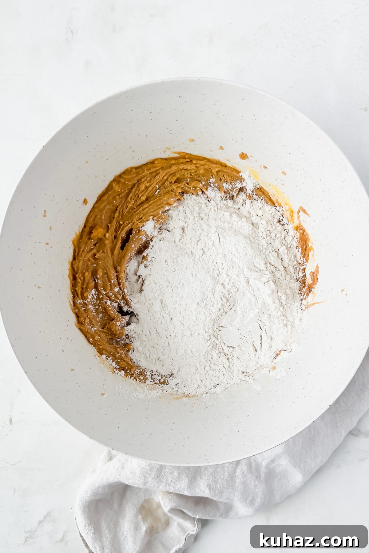 Almond Butter Blossom Cookies 4 wet ingredients for peanut butter crinkle cookies mixed and topped with dry ingredients in white bowl.