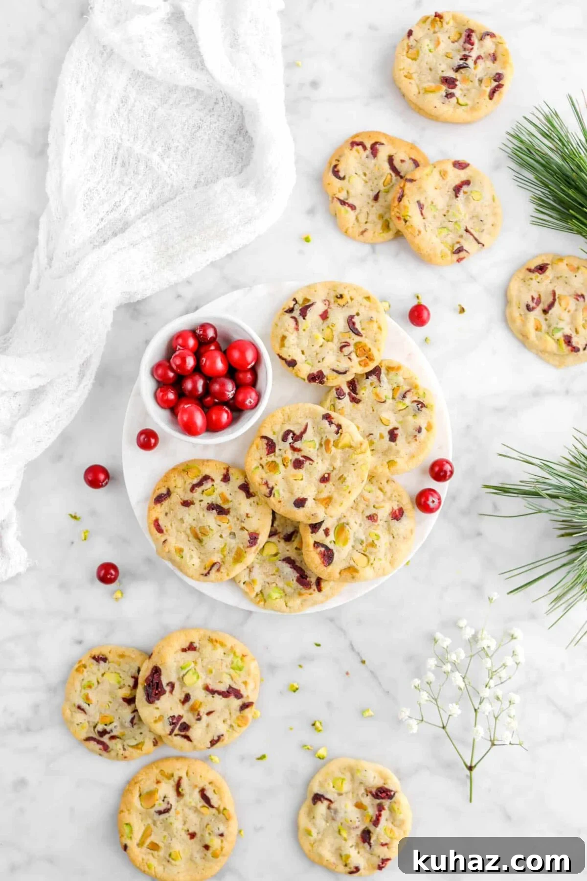 Cranberry Pistachio Slice-and-Bake Cookie Recipe 8 pulled back flat lay of cranberry pistachio cookies with six cookies on round marble plate with small bowl of cranberries, eight more cookies around, flowers, and a napkin