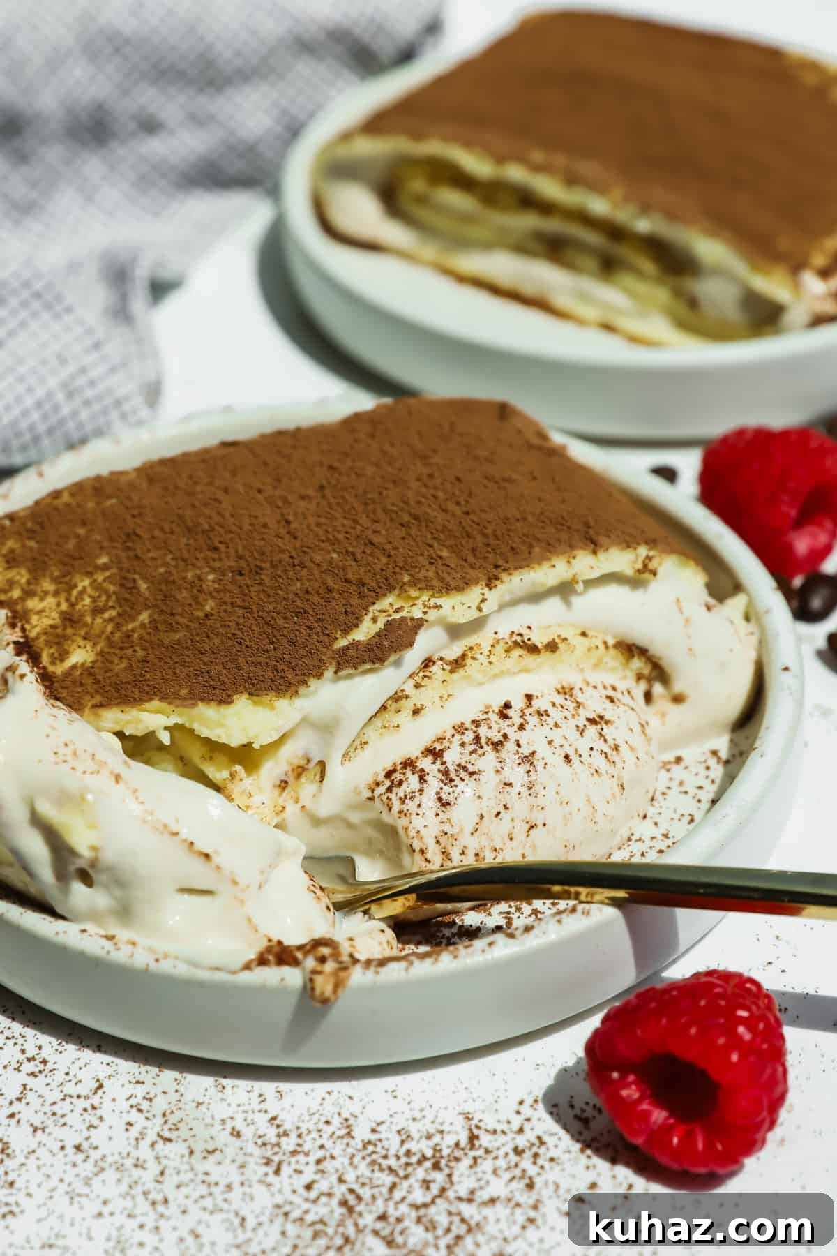 Tiramisu Crepe Roll with Cottage Cheese 2 A plate of cottage cheese tiramisu crepe roll with a portion scooped out reveals creamy layers, dusted with cocoa powder. Fresh raspberries and a gold spoon are nearby.