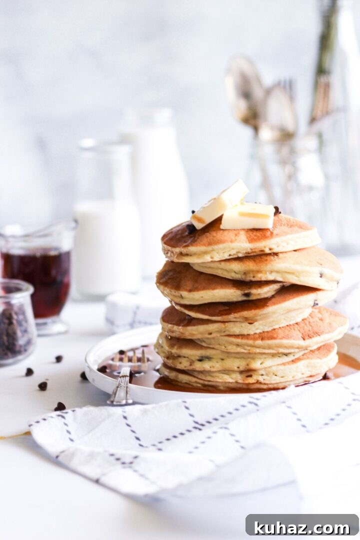 Gluten Free Buttermilk Pancakes Recipe 2 A fluffy stack of pancakes with butter on top served on a white plate with syrup, milk, and chocolate chips nearby.