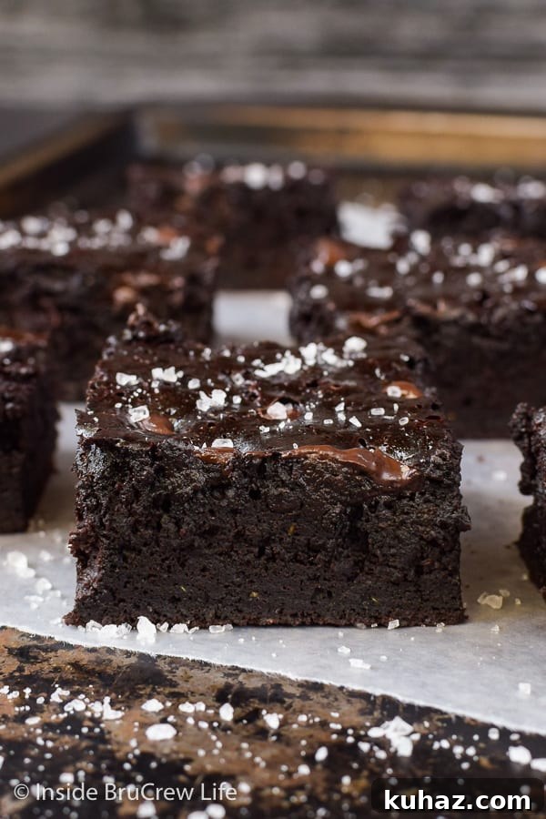 Fudgy Dark Chocolate Zucchini Brownies - shredded zucchini transforms these homemade brownies into a rich and fudgy dessert. Try this easy recipe and enjoy the sweet and salty goodness! Close up picture of a dark chocolate zucchini brownie on a tray with more brownies behind it