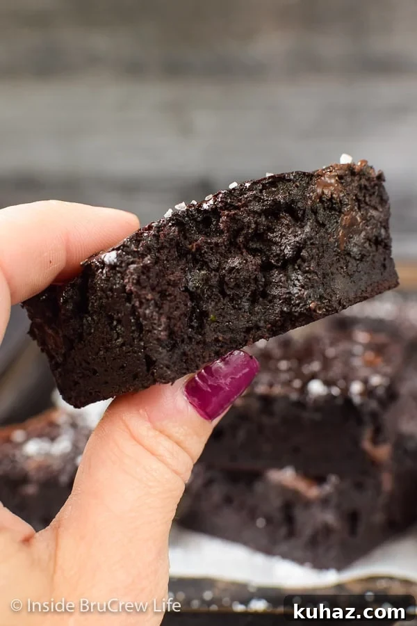 Fudgy Dark Chocolate Zucchini Brownies - these rich and fudgy brownies are actually made with zucchini. The green veggies makes this homemade brownie recipe taste amazing! A hand holding a dark chocolate zucchini brownie up close with a bite out of it