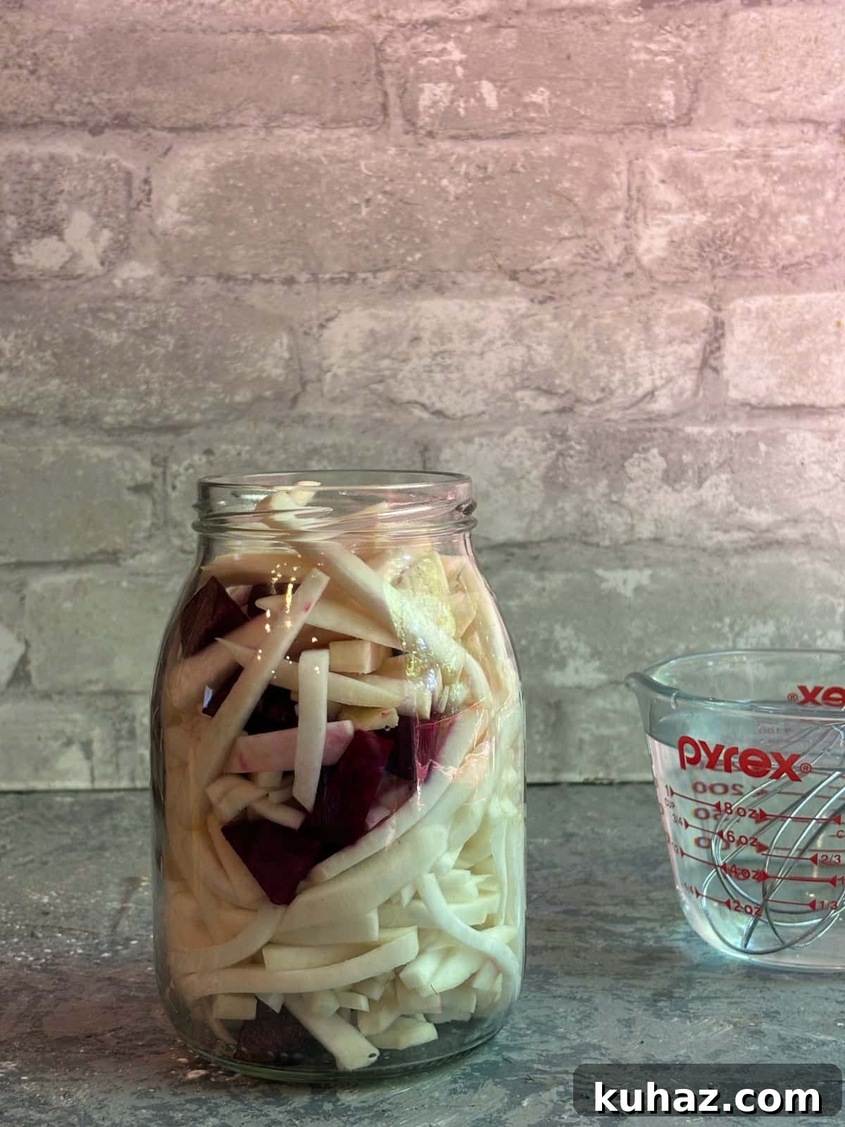 chopped turnips, beets, and garlic in a large glass jar with a liquid measuring cup in the background.