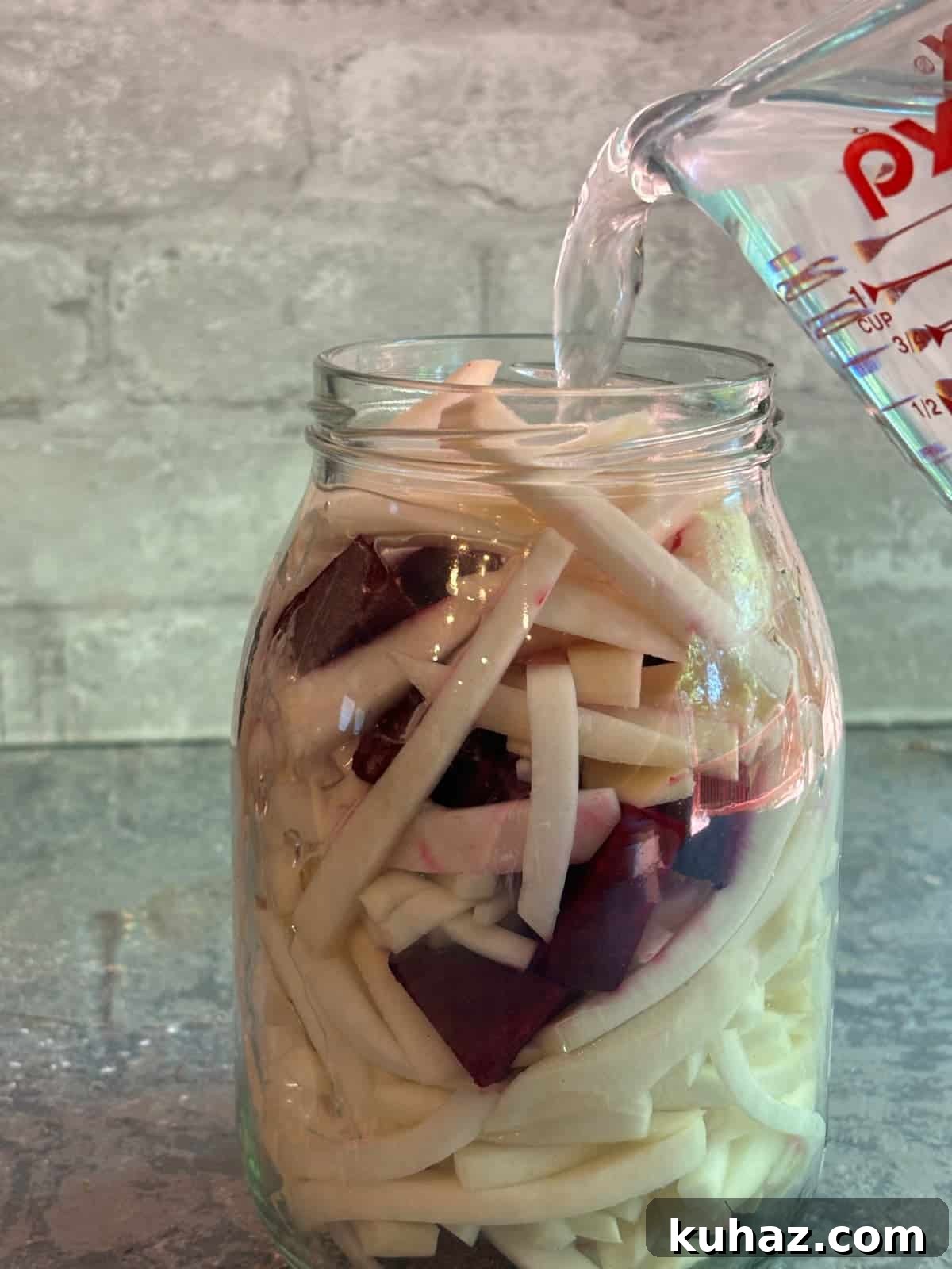 chopped turnips, beets, and garlic in a large glass jar. A liquid measuring cup is pouring clear brining liquid into the glass jar.