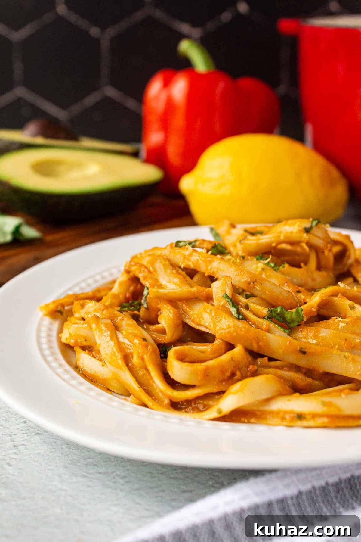 Creamy Avocado Bell Pepper Pasta 2 Avocado bell pepper pasta on a plate. Fettuccine noodles coated in sauce garnished with fresh basil. Avocado, red bell pepper, and lemon are in the background.