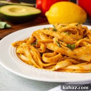 Creamy Avocado Bell Pepper Pasta 11 Avocado bell pepper pasta on a plate. Fettuccine noodles coated in sauce garnished with fresh basil. Avocado, red bell pepper, and lemon are in the background.