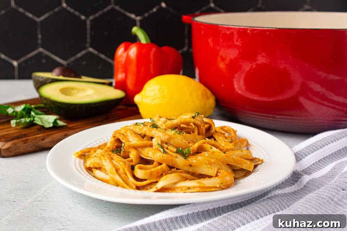 Creamy Avocado Bell Pepper Pasta 9 Fettuccine noodles coated in creamy avocado roasted red pepper sauce garnished with fresh basil and lemon zest. Avocado, red bell pepper, basil, and lemon are in the background.