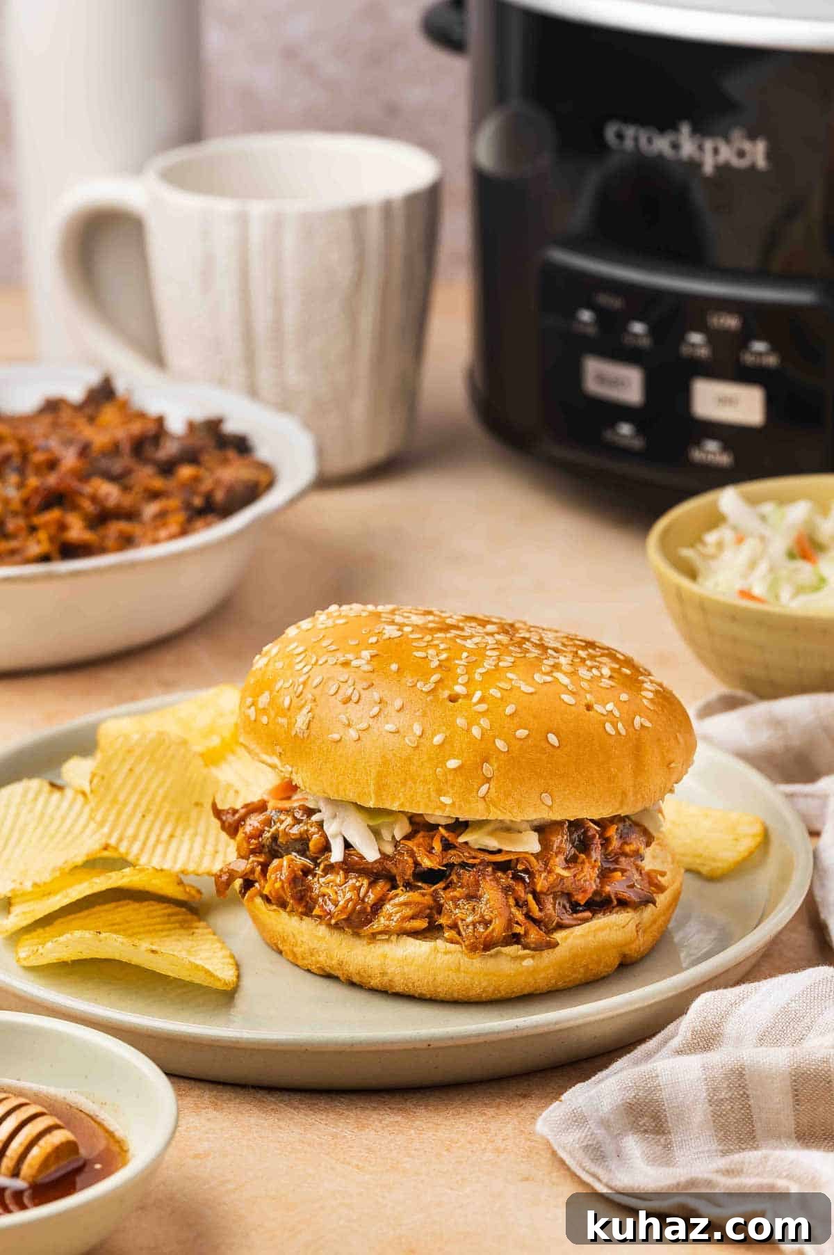 A plate with shredded slow cooker hot honey chicken inbetween burger buns with some chips on the side and a slow cooker in the background.