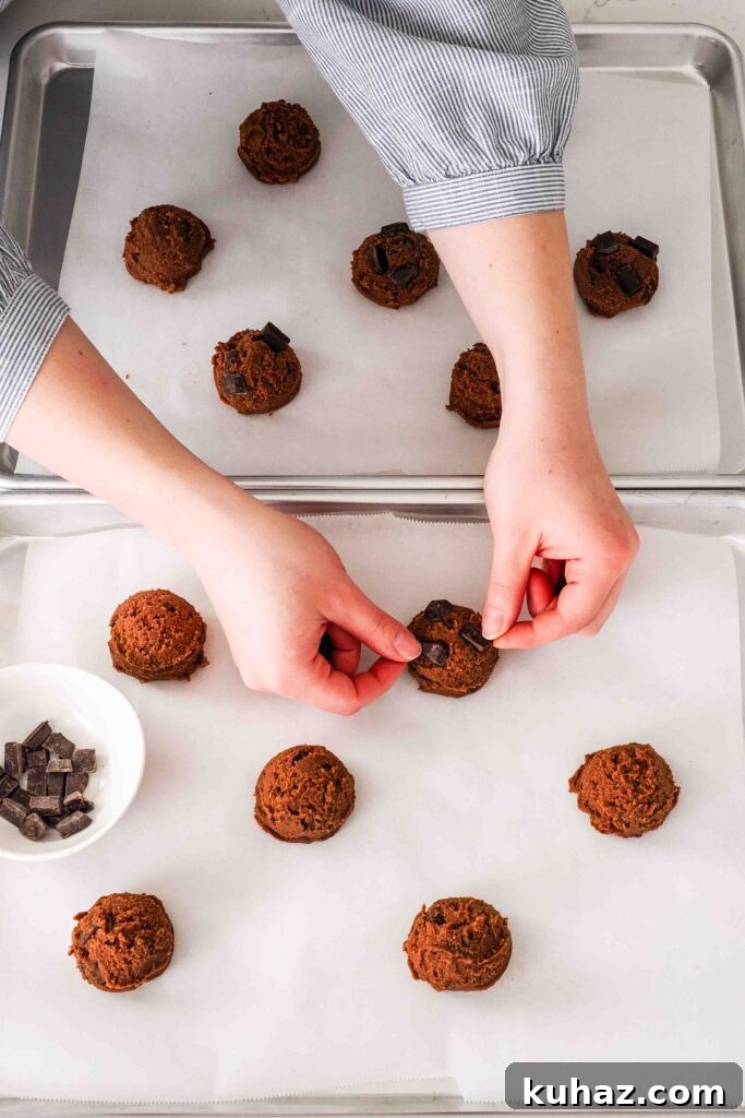 Chewy Double Chocolate Chip Cookies Recipe 11 Two hands push more chocolate chips onto cookie dough balls on baking sheets.