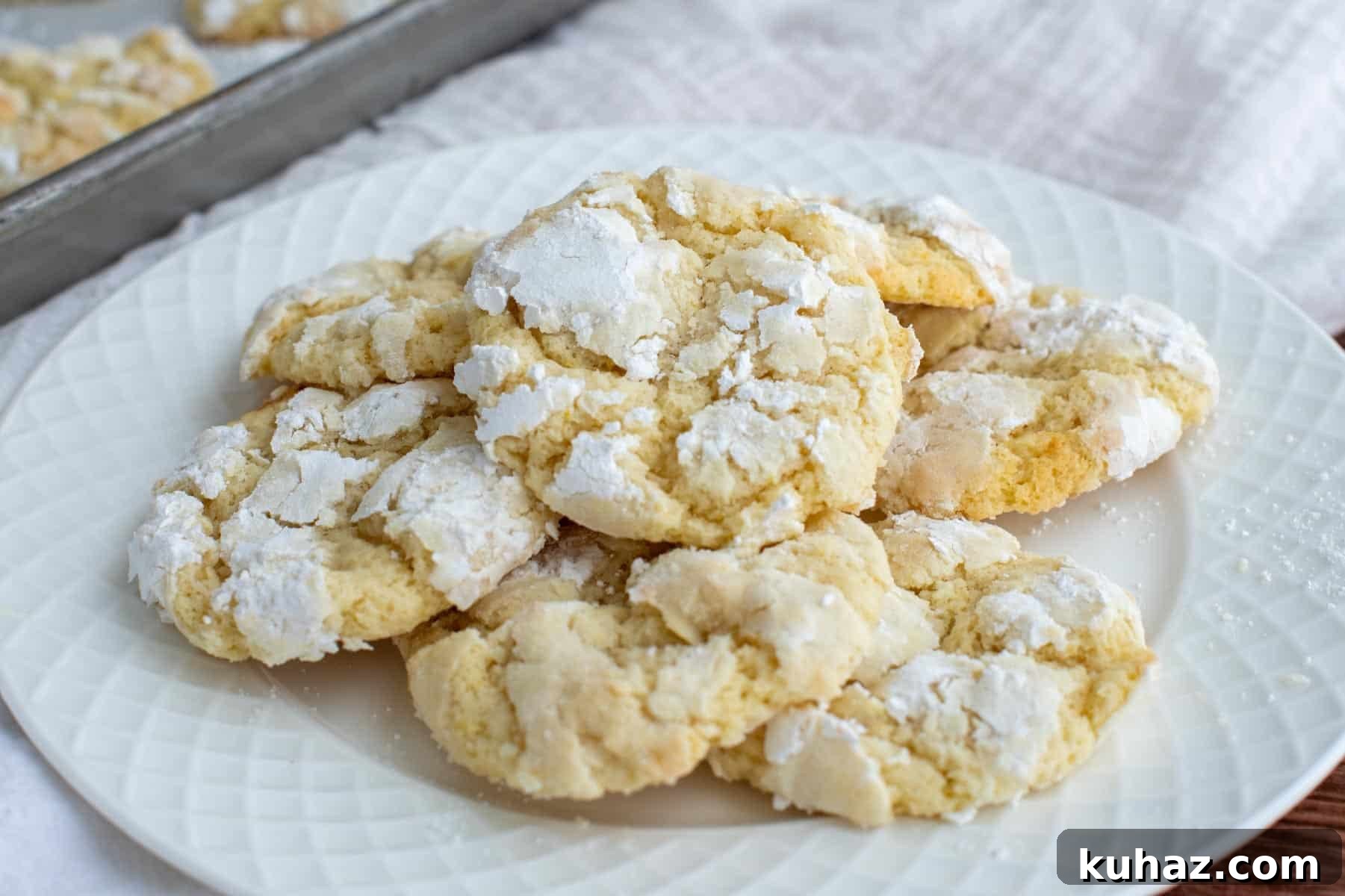 Lemon Crinkle Cookies Made with Sourdough Discard 2 Pile of lemon crinkle cookies with powdered sugar coating and crackled tops on a white plate, with more cookies on a baking sheet in the background.
