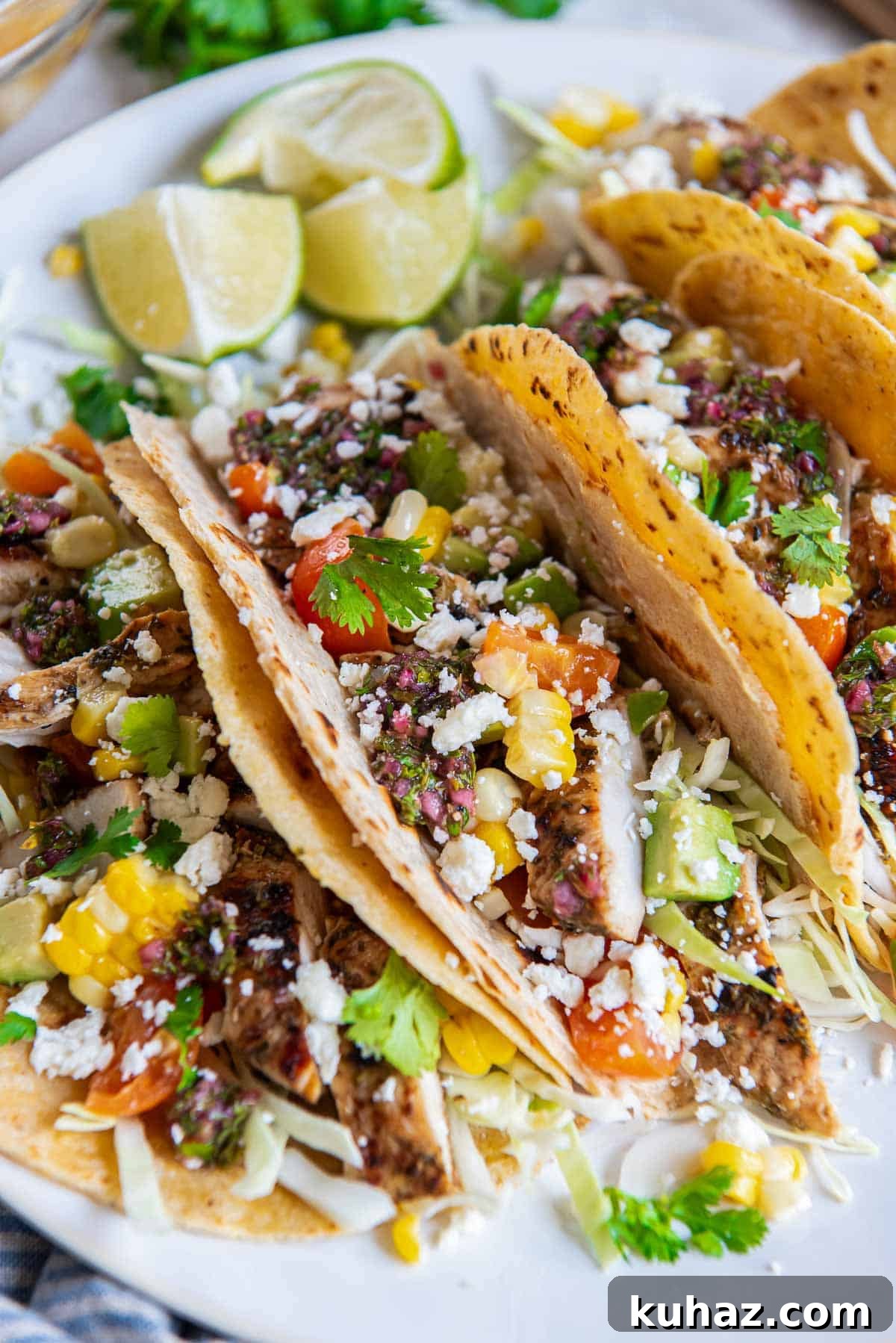 Grilled Chimichurri Chicken Tacos with Lime & Cilantro 3 Three chimichurri chicken tacos topped with corn, tomatoes, and cabbage on a platter with lime wedges.