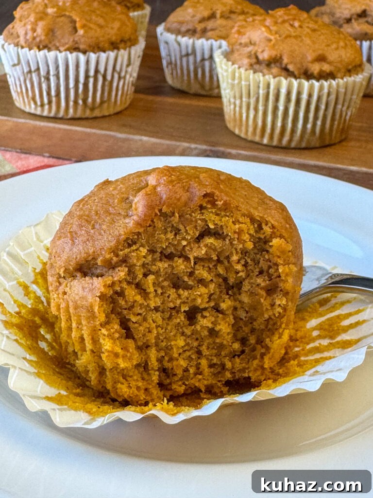 Pumpkin Spice Cupcake Recipe with Cream Cheese Frosting 9 Bite taken out of a delightful pumpkin spice cupcake that's returned to rest on a liner on a white plate with a fork and the rest of the cupcakes in the recipe in the background.