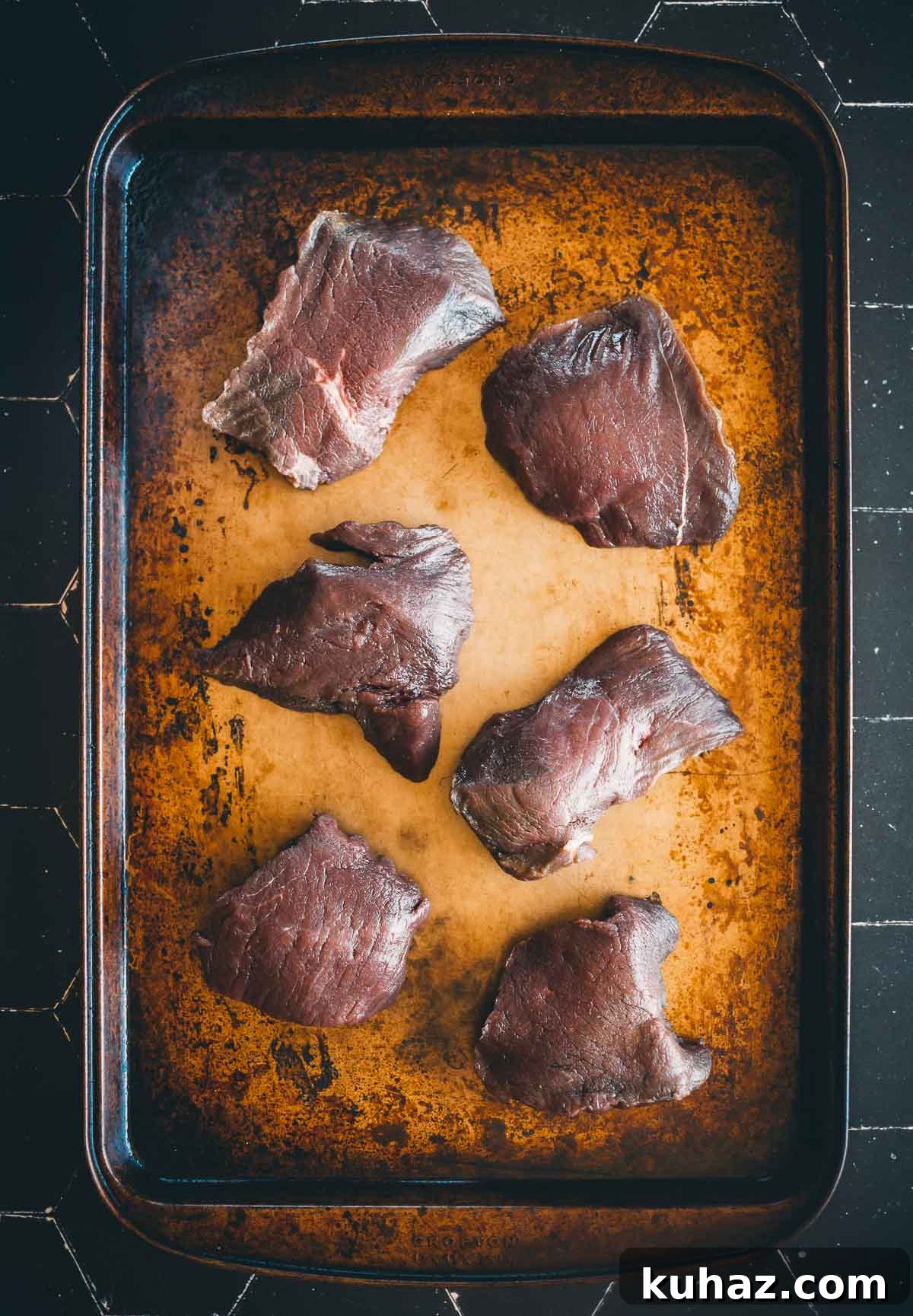 How to Grill Tender Elk Steaks with Smoky Flavor 3 Six raw elk steaks displayed on a well-used, darkened baking sheet.