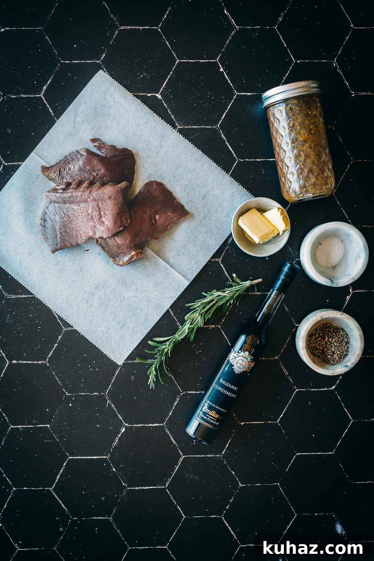 How to Grill Tender Elk Steaks with Smoky Flavor 4 Raw elk steaks on a parchment paper, jar of seasoning, bowl of butter, rosemary, balsamic vinegar bottle, and bowls of salt and pepper placed on a black tiled surface.