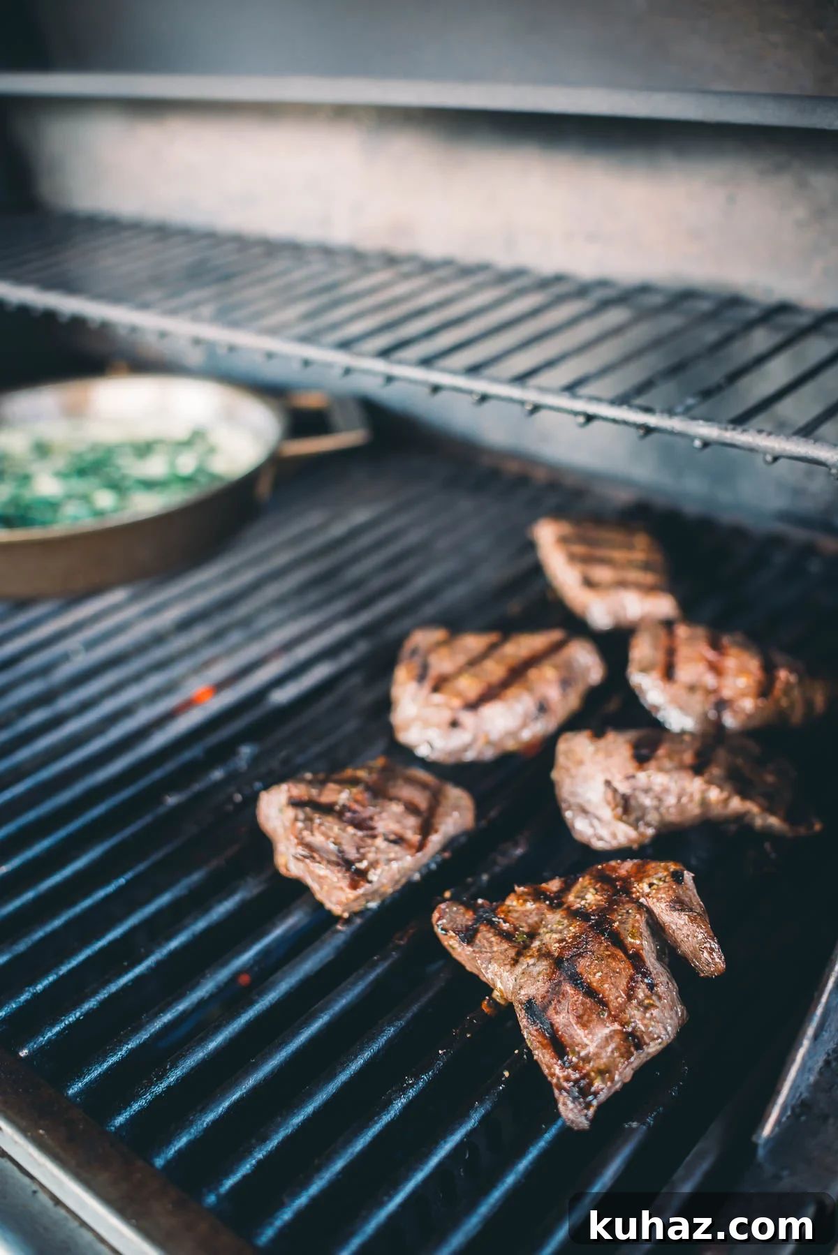 How to Grill Tender Elk Steaks with Smoky Flavor 7 Several elk steaks are grilling on a barbecue. A skillet with vegetables is also visible in the background.