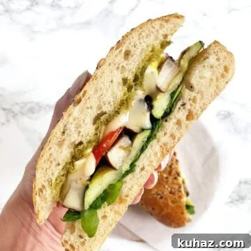 A hand holding up a sandwich with roasted vegetables, brie, and pesto