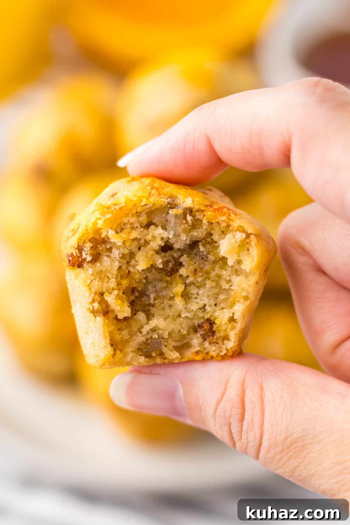 Savory Breakfast Sausage Pancake Muffins 3 A hand holding a sausage McGriddle bite with a bite taken out, revealing a fluffy interior with pieces of sausage. The background shows more sausage pancake muffins stacked on a plate.
