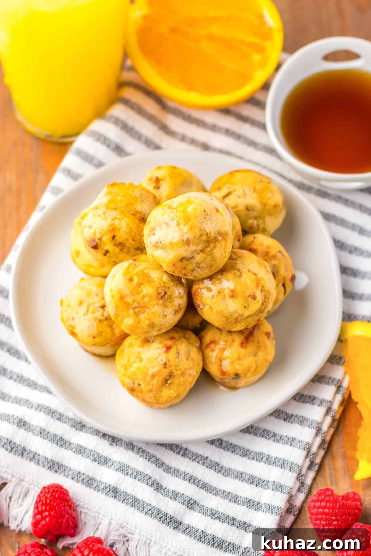 Savory Breakfast Sausage Pancake Muffins 7 A plate of golden-brown sausage pancake muffins stacked high, drizzled with syrup. The plate sits on a striped cloth napkin with fresh raspberries, orange slices, a cup of syrup, and a glass of orange juice in the background.