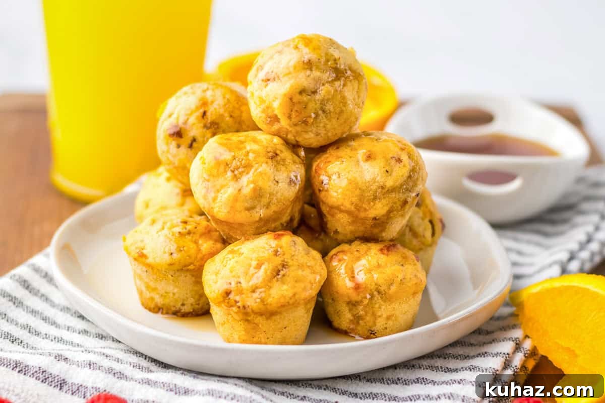 Savory Breakfast Sausage Pancake Muffins 8 A stack of mini sausage and pancake muffins arranged on a white plate, drizzled with syrup. A glass of orange juice, a cup of syrup, and orange slices are in the background.