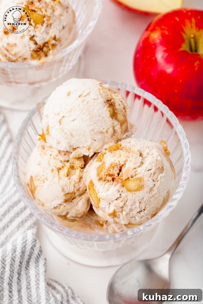 overhead shot of a glass ice cream dish with three scoops of homemade apple pie ice cream. Next to the dish is a silver spoon and a red apple
