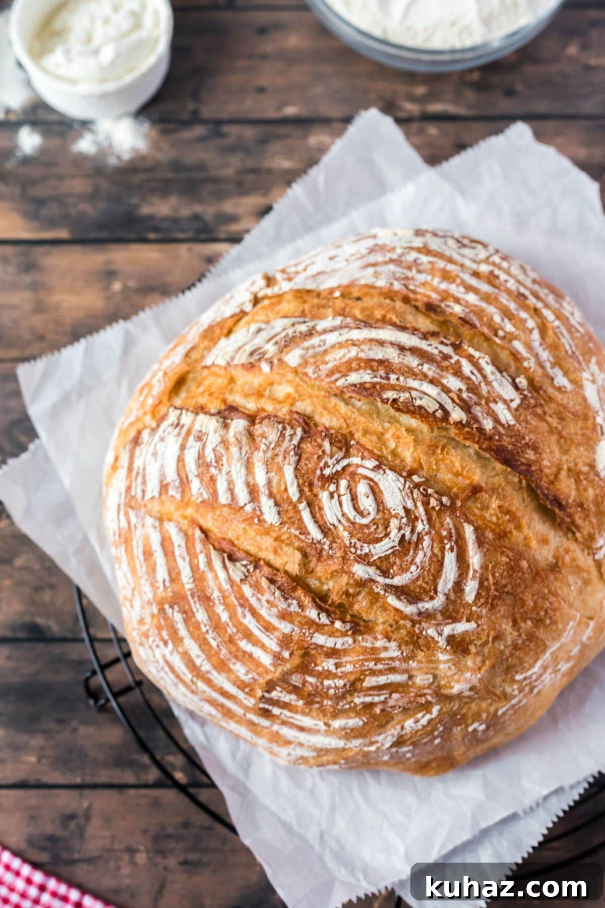 A round loaf of sourdough bread on top of white parchment paper laying on a wood table with flour on the side.