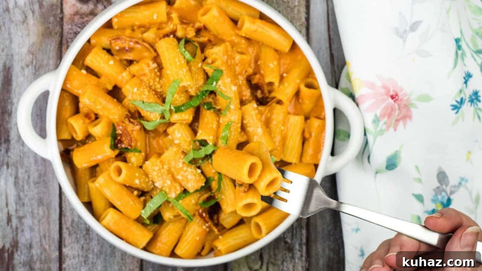17 Authentic Italian Dinner Recipes Even Nonna Will Love 19 A white bowl of rigatoni pasta with creamy sauce, garnished with cheese and green herbs, is placed on a wooden surface. A hand is holding a fork with a piece of pasta.