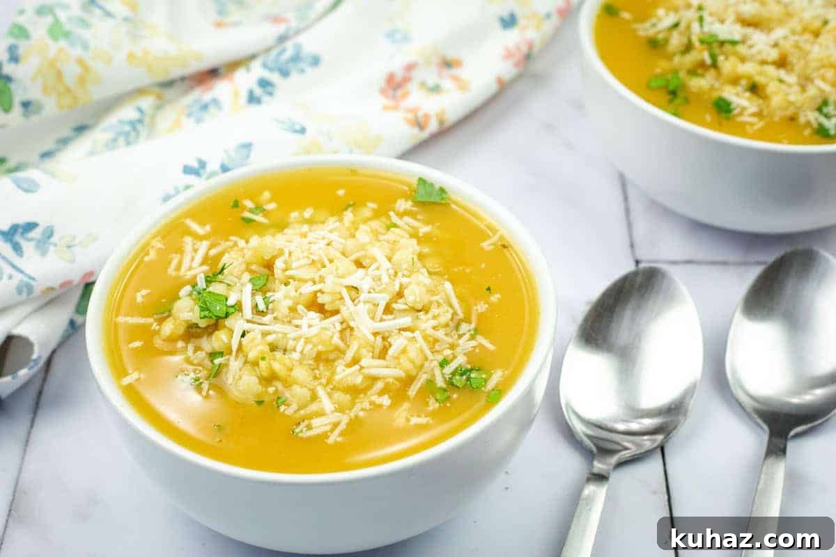 17 Authentic Italian Dinner Recipes Even Nonna Will Love 4 A bowl of Nonna’s Pastina Soup topped with shredded cheese and herbs, next to two silver spoons on a marble surface with a floral cloth in the background.