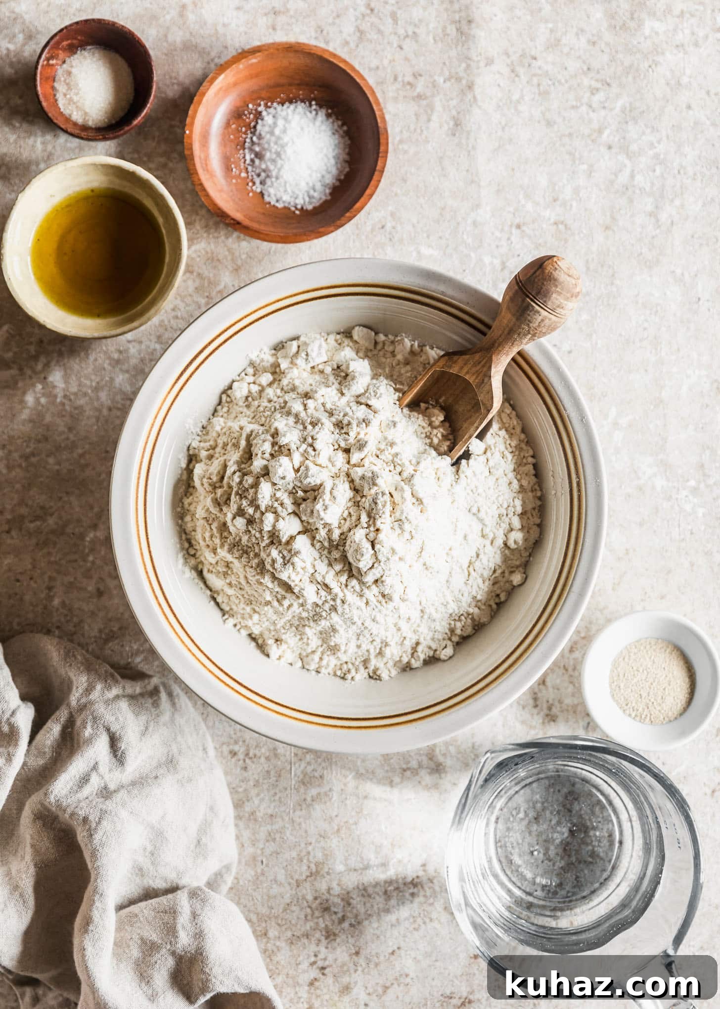Sicilian-Style Pizza: High-Altitude Baking Tips 3 A white bowl of flour next to white bowls of yeast and olive oil, wood bowls of salt and sugar, a pitcher of water, and a beige linen on a tan counter.