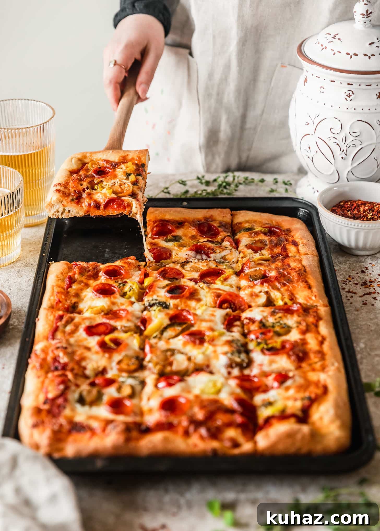 Sicilian-Style Pizza: High-Altitude Baking Tips 7 A woman wearing a tan apron using a wood spatula to pick up a piece of Sicilian-style pizza off of a sheet pan on a tan counter next to glasses of beer, thyme, a white bowl of red pepper flakes, and a white canister.
